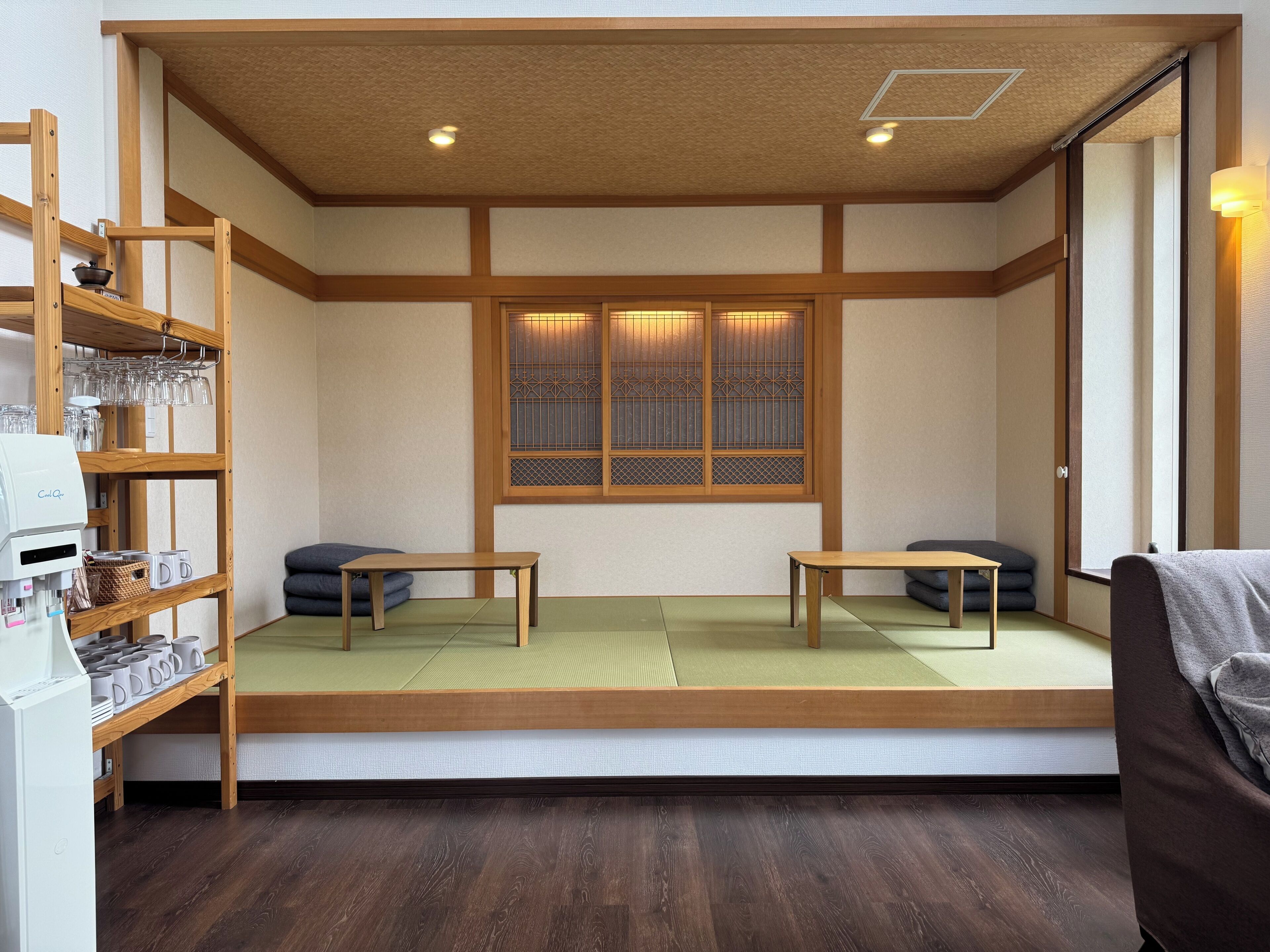 A traditional Japanese room with tatami flooring, a low wooden bed, a small table, a shelf with glasses and cups, and a window with a decorative screen.