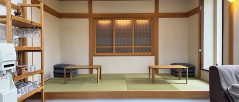 A traditional Japanese room with tatami flooring, a low wooden bed, a small table, a shelf with glasses and cups, and a window with a decorative screen.