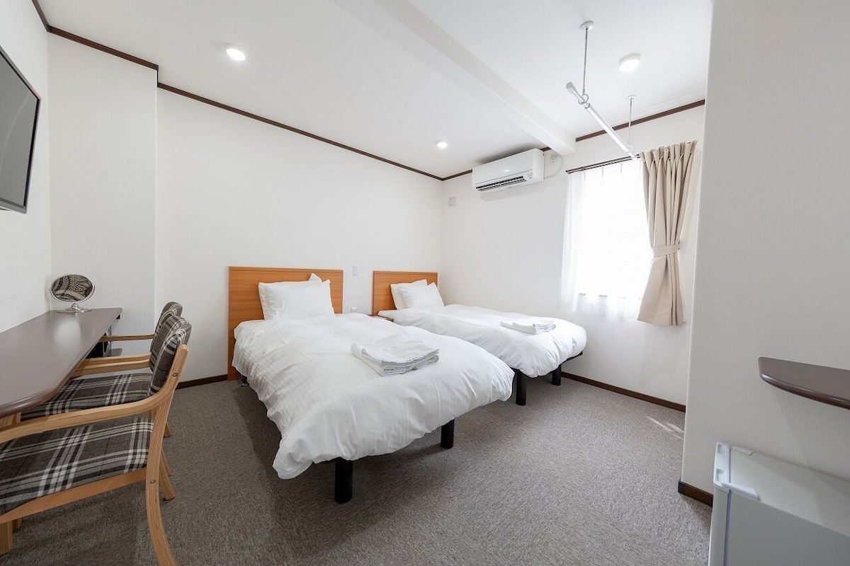A hotel room with two beds, a desk, a chair, an air conditioning unit, and a window with curtains.