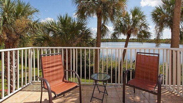 A balcony with two wicker chairs and a small glass table, overlooking a body of water and palm trees. - LMI Gulf Coast Homes, Port Charlotte (Port Charlotte)