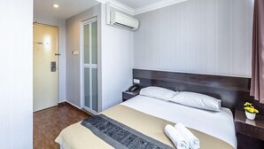 A hotel room with a bed, a nightstand, an air conditioning unit, a bathroom with a glass door, and a small table with flowers.