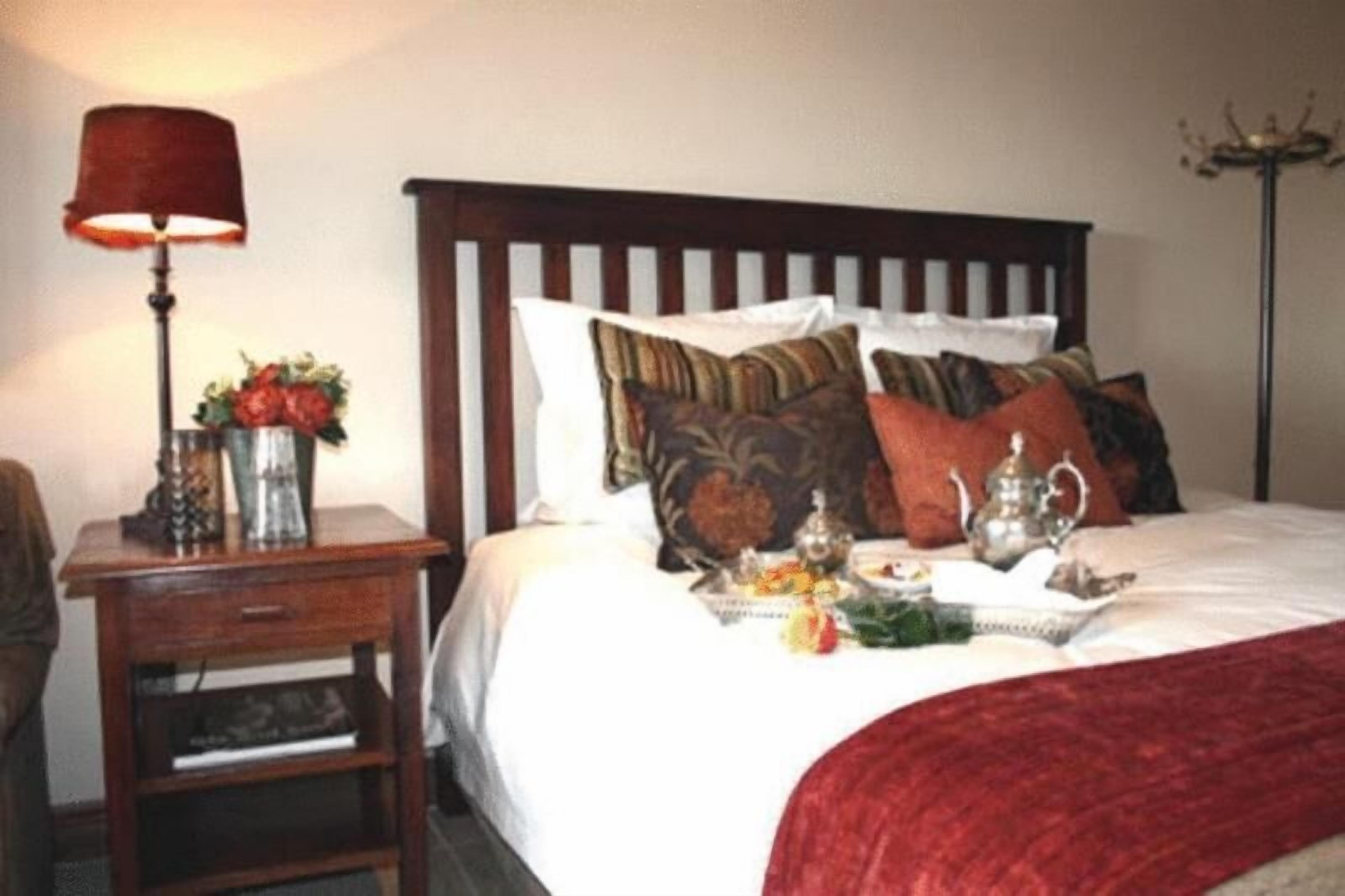 Un lit bien fait, recouvert d’une couverture rouge, une table de chevet avec une lampe, et un plateau avec du thé et des fleurs.