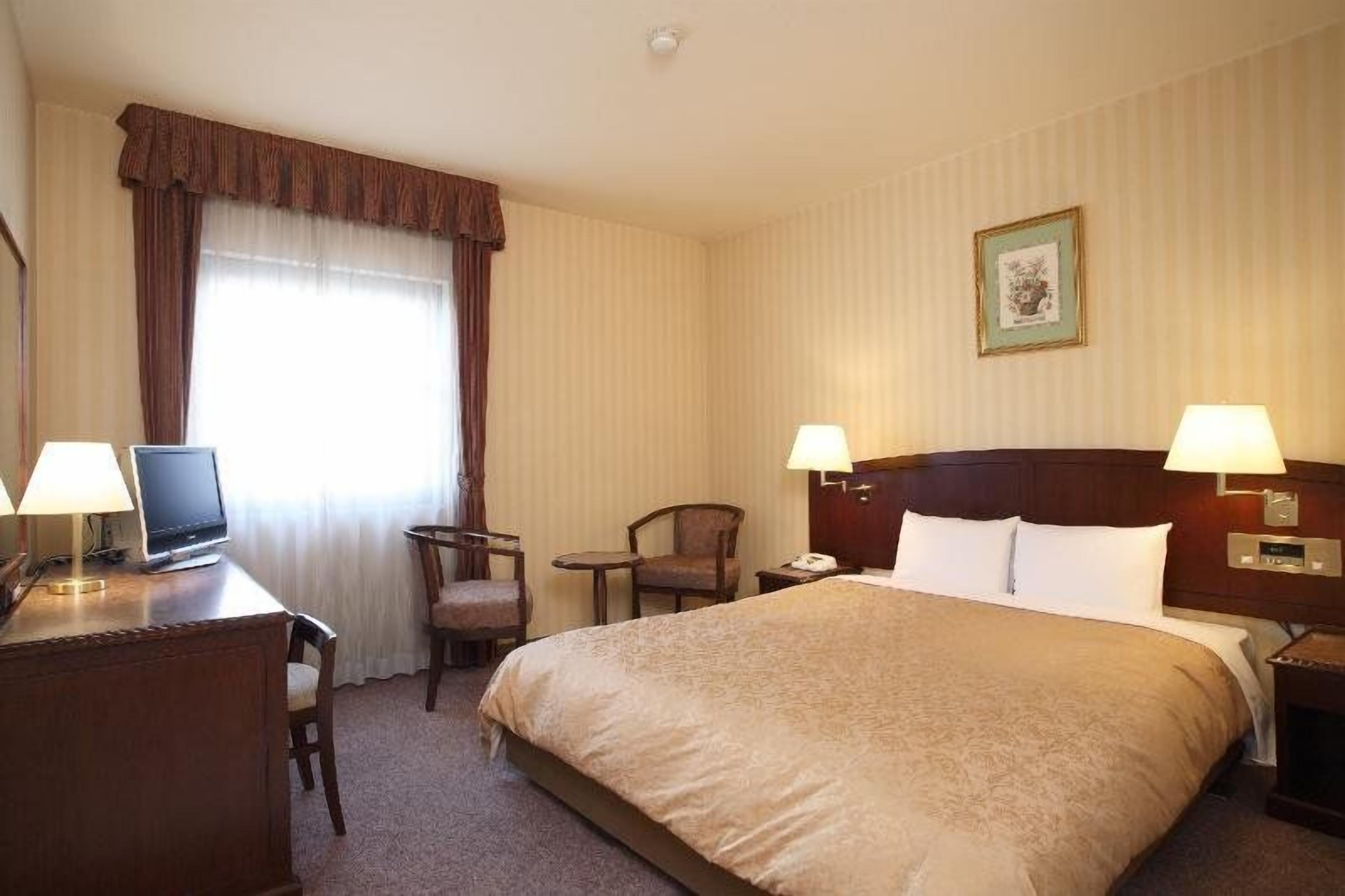 Une chambre d’hôtel avec un grand lit, deux lampes de chevet, un bureau avec un ordinateur, une chaise et une petite table.
