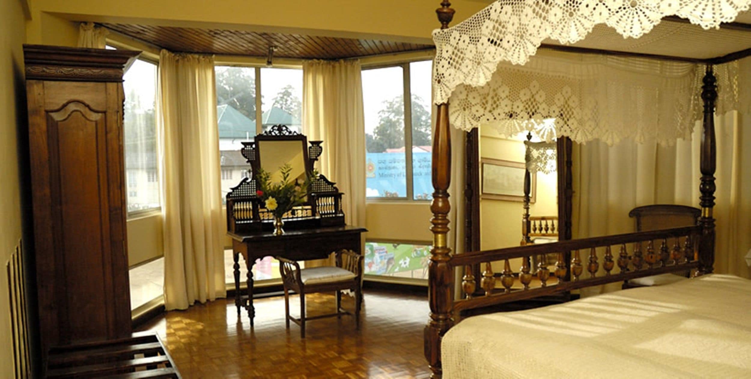 A four-poster bed with a wooden headboard, a wooden desk with a mirror and a vase of flowers, and a view of the outdoors through large windows.