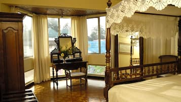 A four-poster bed with a wooden headboard, a wooden desk with a mirror and a vase of flowers, and a view of the outdoors through large windows.
