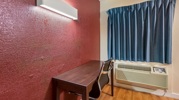 A room with a wooden table, a chair, a wall-mounted air conditioner, and a window with blue curtains. - Red Roof Inn Plano (Plano)