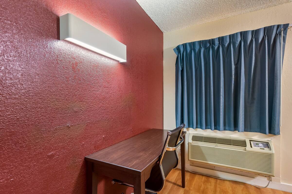 A room with a wooden table, a chair, a wall-mounted air conditioner, and a window with blue curtains.