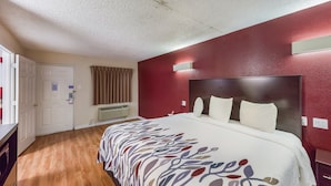 A hotel room with a wooden floor, a large bed with a patterned bedspread, a red accent wall, and a white door. - Red Roof Inn Plano (Plano)