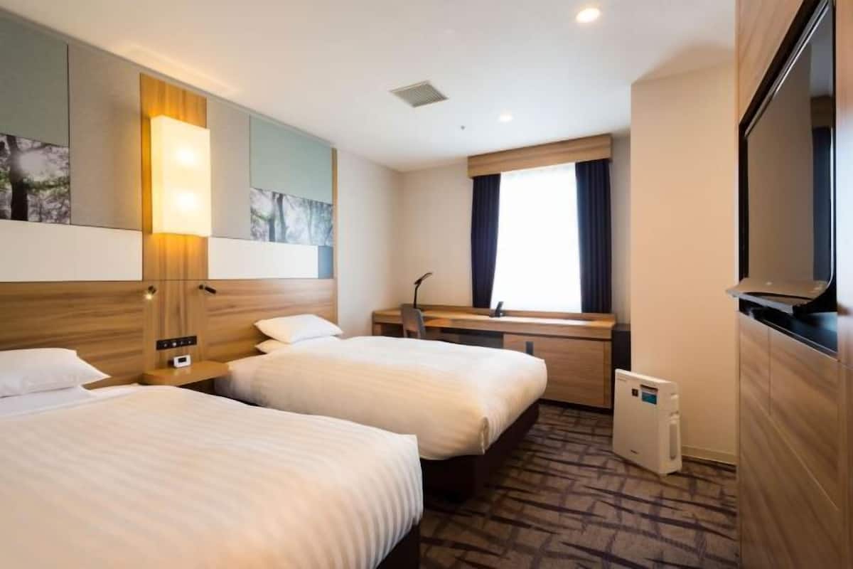Quarto de hotel com duas camas, uma escrivaninha, televisão e janela com cortinas.