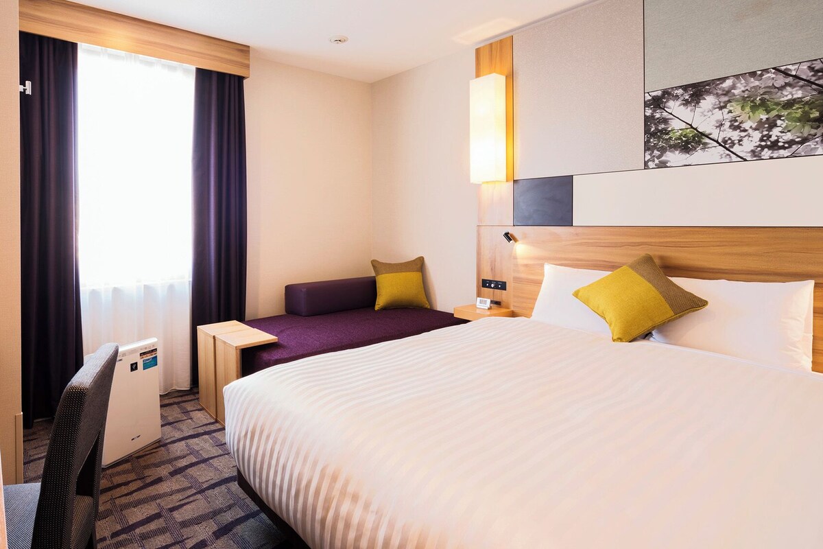 Habitación de hotel con cama, escritorio, silla, mesita y ventana con cortinas.