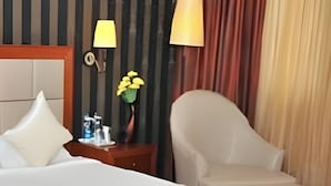 Una habitación de hotel con cama, una silla, una mesita de noche con lámpara y un florero con flores.