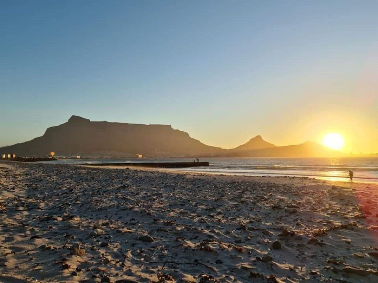 A beach with a pier extending into the water, a mountain in the background, and the sun setting over the horizon.
