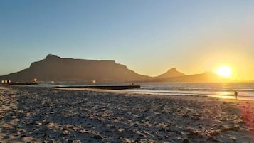 A beach with a pier extending into the water, a mountain in the background, and the sun setting over the horizon.