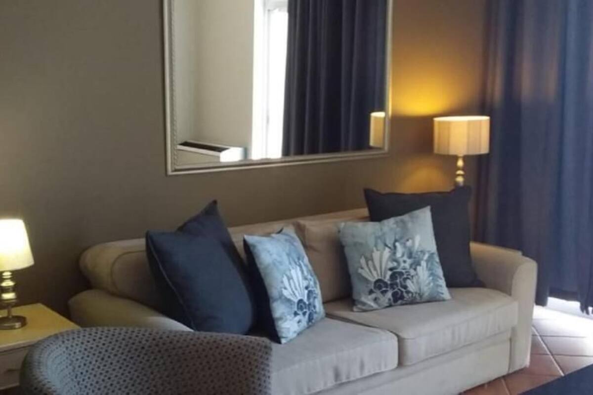A hotel room with a sofa, two pillows, a side table with a lamp, and a large mirror on the wall.