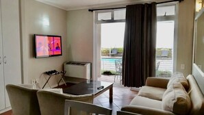 A living room with a TV, a dining table, chairs, a sofa, and a view of a balcony with a pool. - Leisure Bay (Cape Town)