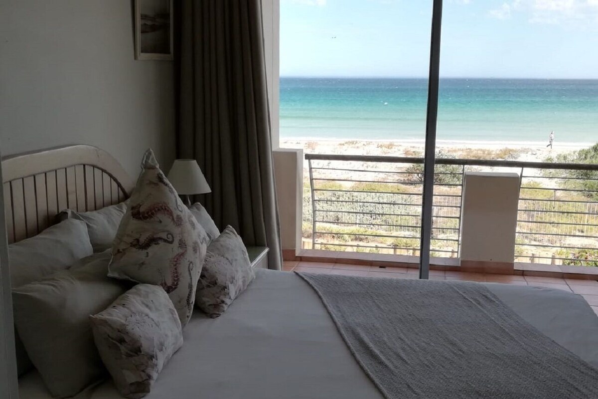 A bedroom with a bed, pillows, a nightstand, a lamp, and a view of the beach through a balcony.