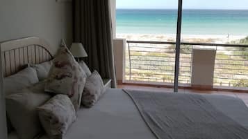 A bedroom with a bed, pillows, a nightstand, a lamp, and a view of the beach through a balcony.