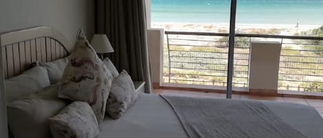 A bedroom with a bed, pillows, a nightstand, a lamp, and a view of the beach through a balcony.