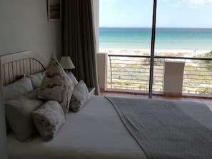 A bedroom with a bed, pillows, a nightstand, a lamp, and a view of the beach through a balcony. - Leisure Bay (Cape Town)