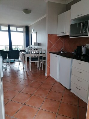 A compact kitchen with white cabinets, a black countertop, and a microwave. There is a dining area with a table and chairs, and a window with curtains. - Leisure Bay (Cape Town)