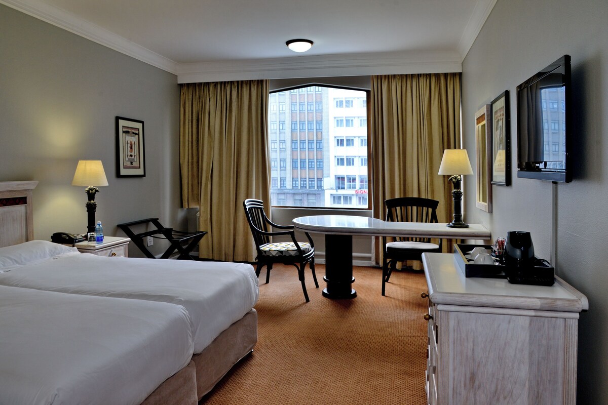 A hotel room with two beds, a desk, a chair, a lamp, a TV, and a window with curtains.