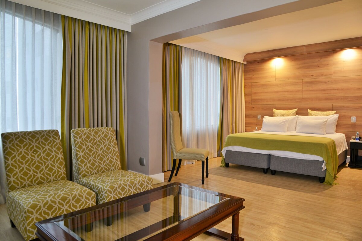 A hotel room with a bed, two chairs, a glass-top table, and curtains.