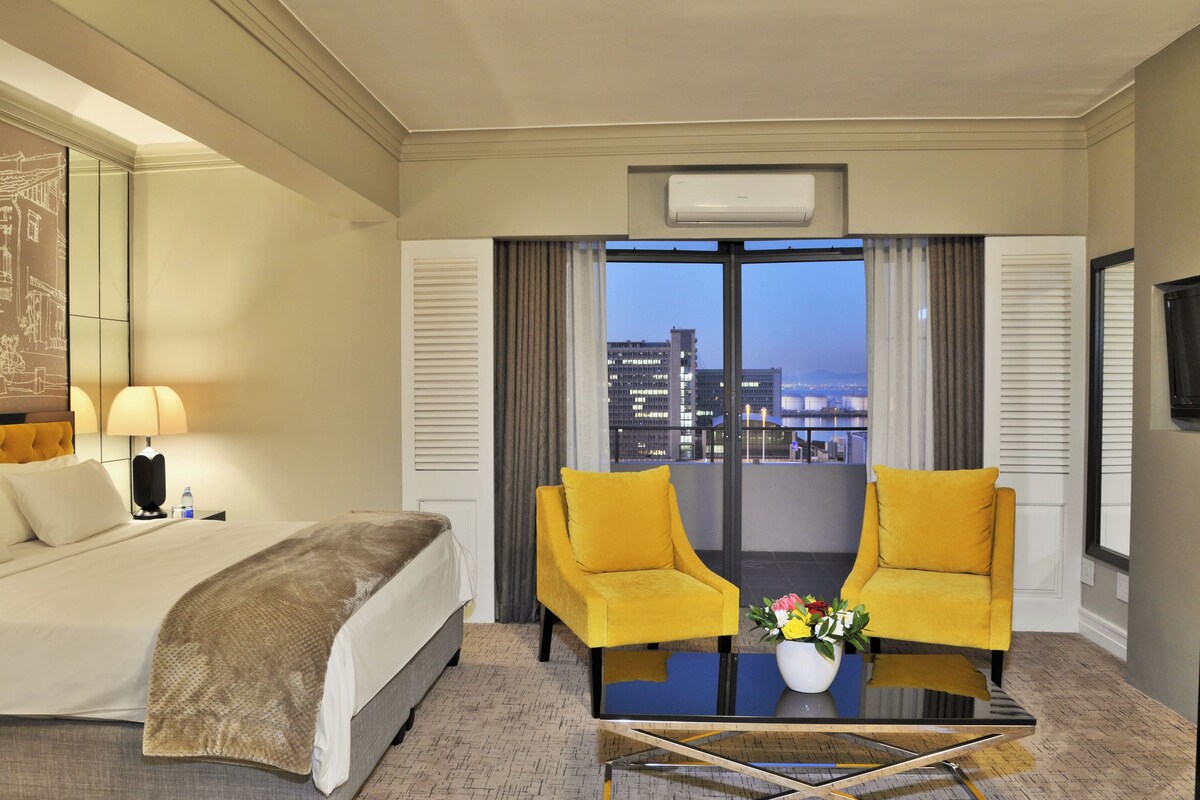 A hotel room with a bed, two yellow armchairs, a coffee table with flowers, a large window with a city view, and an air conditioning unit.