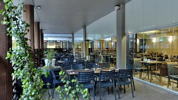 Ein Essbereich mit Tischen und Stühlen, einer Kübelpflanze und Blick auf das Restaurant durch Glaswände.