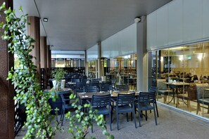 Een overdekte eetruimte met tafels en stoelen, een potplant en uitzicht op het restaurant door glazen wanden.