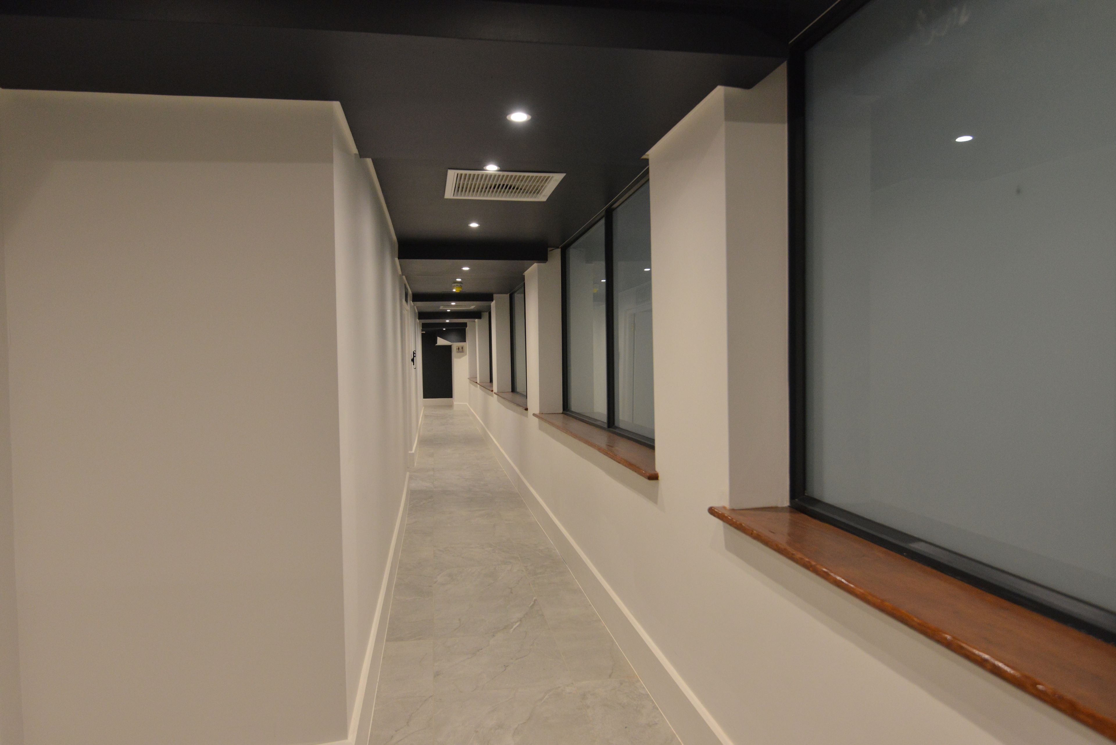 A long, narrow hallway with a tiled floor, recessed lighting, and large windows on one side.