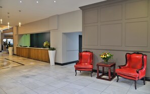 A hotel lobby with a reception desk, two red leather chairs, a small table with a vase of flowers, and a tiled floor. - Capetonian Hotel (Cape Town)