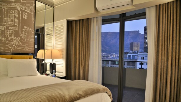 A hotel room with a large bed, a bedside table with a lamp, a window with curtains, and a view of a cityscape and mountains. - Capetonian Hotel (Cape Town)