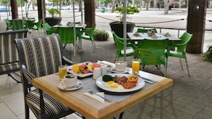 An outdoor dining area with green chairs and tables, a breakfast spread including eggs, beans, and toast, and a view of the street and trees through large windows. - Capetonian Hotel (Cape Town)