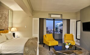 A hotel room with a bed, two yellow armchairs, a coffee table, a TV, and a view of buildings. - Capetonian Hotel (Cape Town)