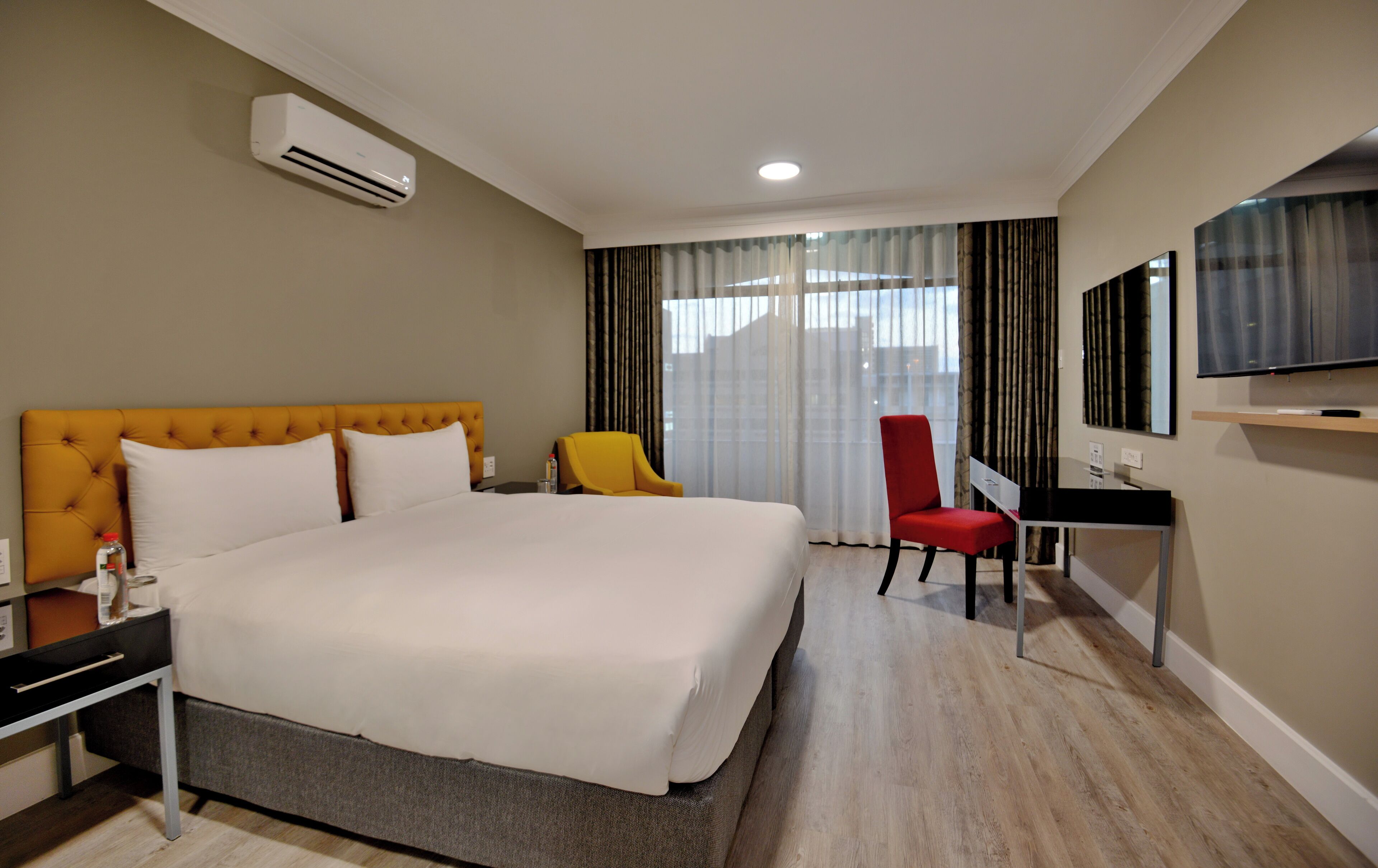 A hotel room with a large bed, a desk, a chair, an air conditioning unit, and a television on the wall.