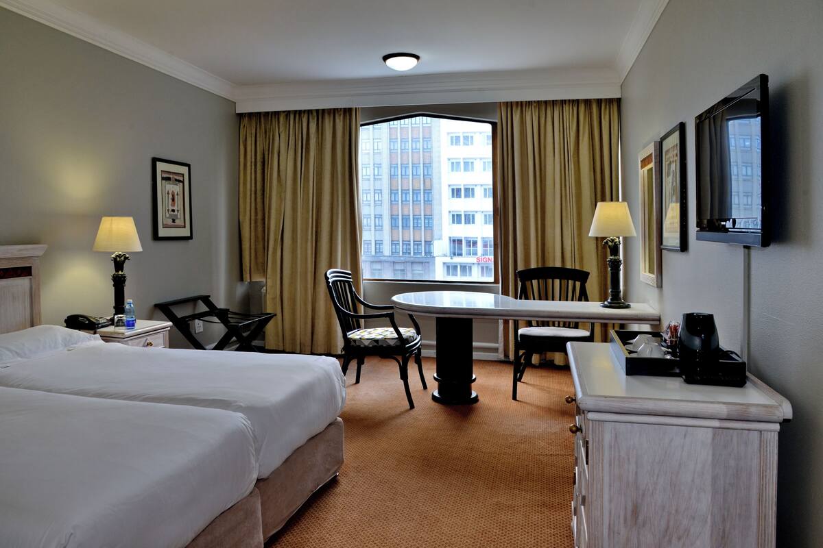 A hotel room with two beds, a desk, a chair, a lamp, a TV, and a window with curtains.