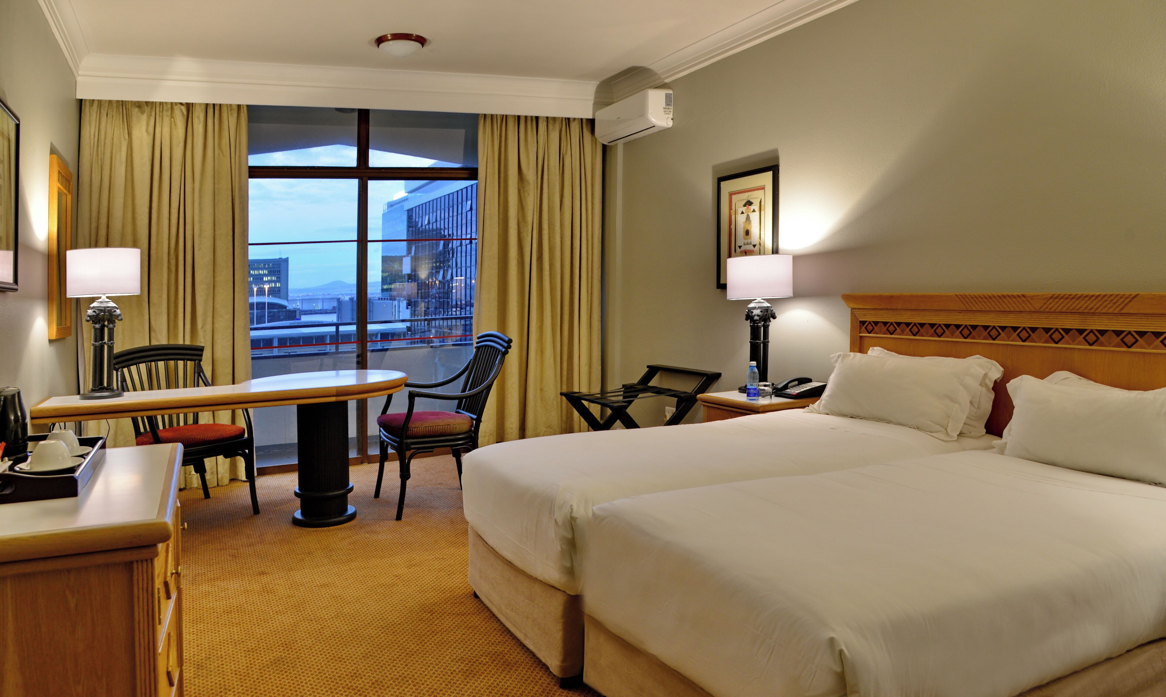 A hotel room with a bed, a desk, a chair, a table, a lamp, a water bottle, a phone, and a window with curtains.