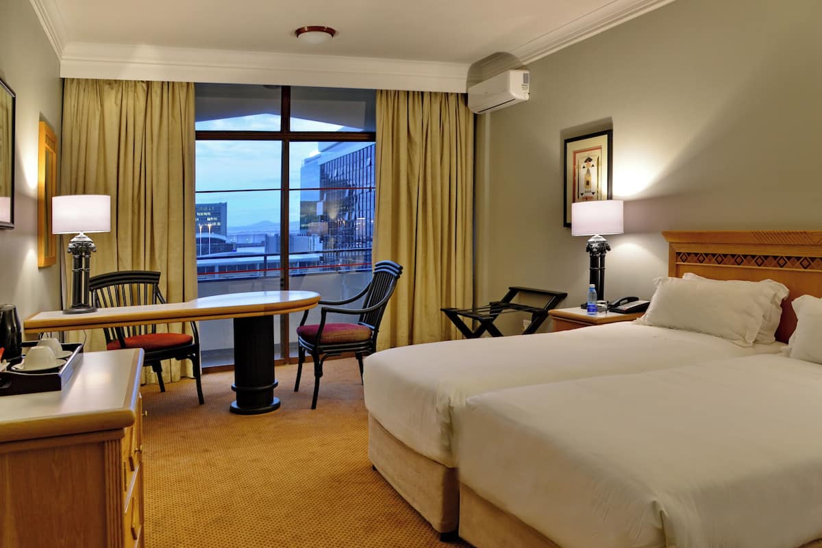 A hotel room with a bed, a desk, a chair, a table, a lamp, a water bottle, a phone, and a window with curtains.