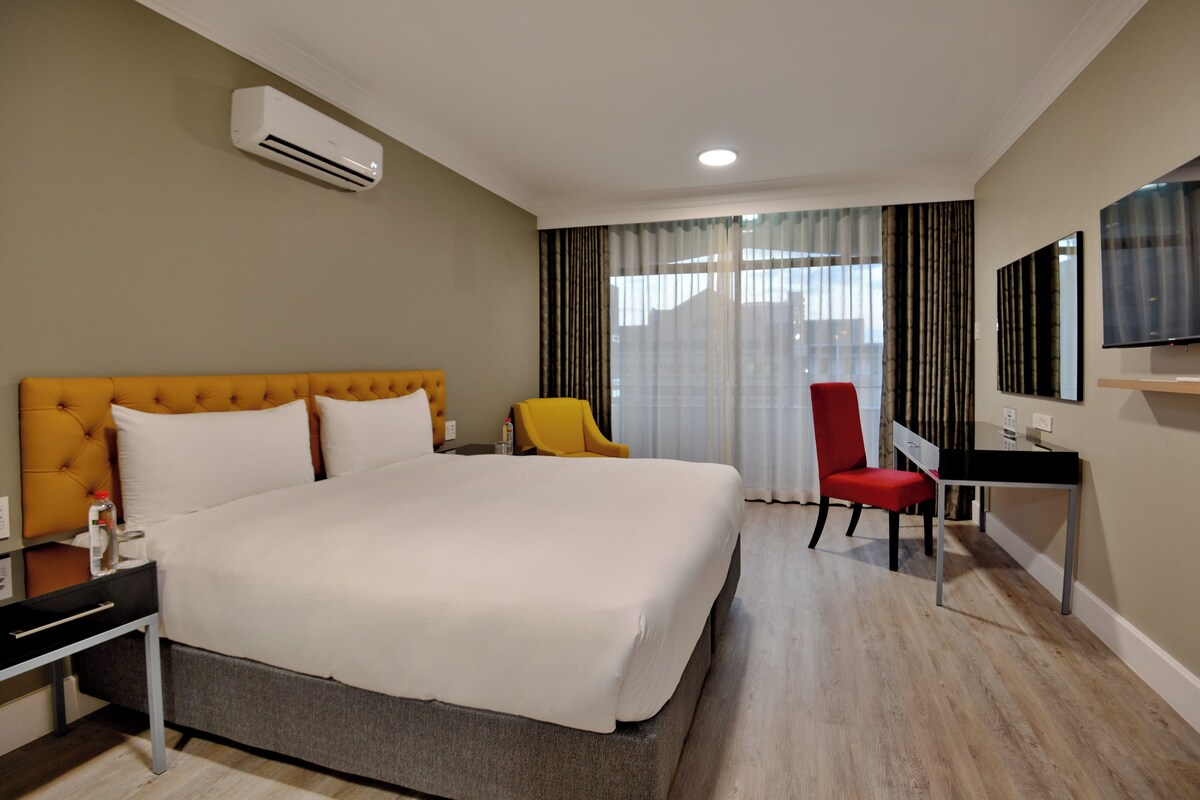 A hotel room with a large bed, a desk, a chair, an air conditioning unit, and a television on the wall.