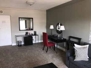 Una habitación con un escritorio, una silla roja, un sofá negro, una lámpara negra y un espejo instalado en la pared.