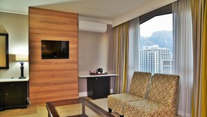 A hotel room with a wooden accent wall, a flat-screen TV, a small desk with a coffee maker, a patterned armchair, and a large window with curtains. - Capetonian Hotel (Cape Town)