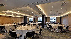 A conference room with multiple round tables, each set with plates, cutlery, and glasses. The room has a modern ceiling design with recessed lighting and large windows offering a view of the outside. - Capetonian Hotel (Cape Town)