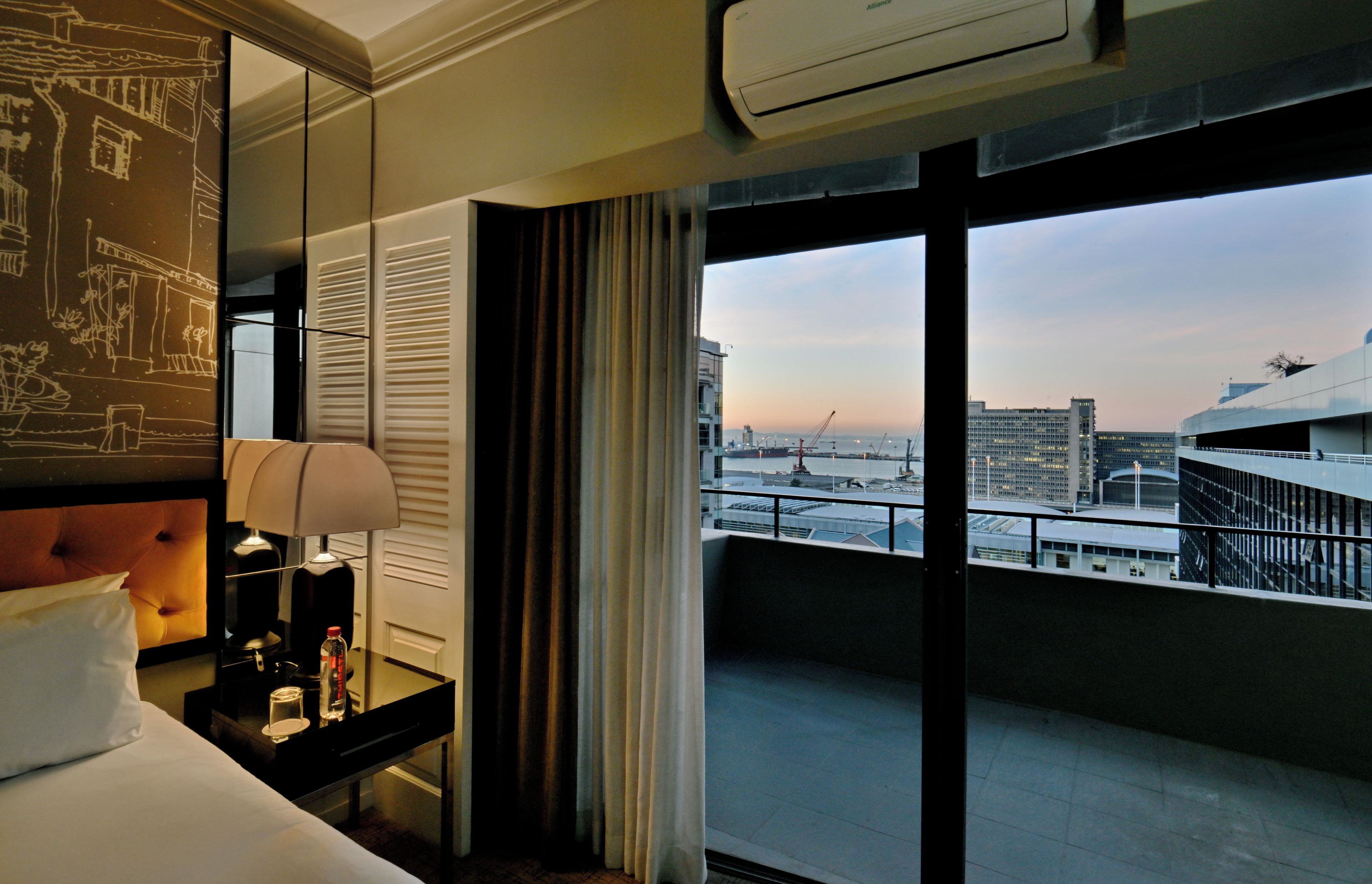 A hotel room with a large window, a bed, a nightstand, and a view of the cityscape.
