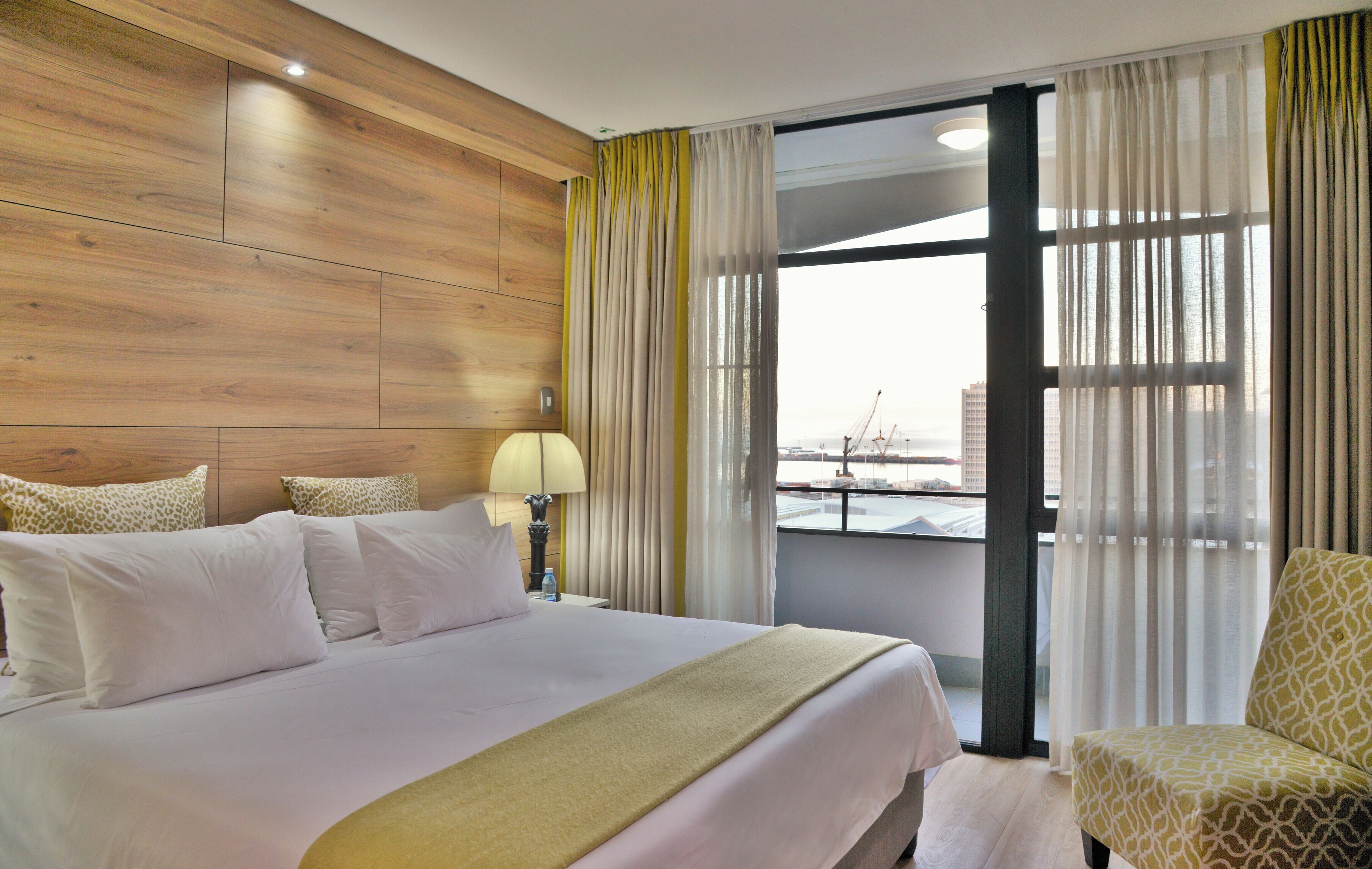 A hotel room with a large bed, a chair, a nightstand with a lamp, and a view of the city through the window.