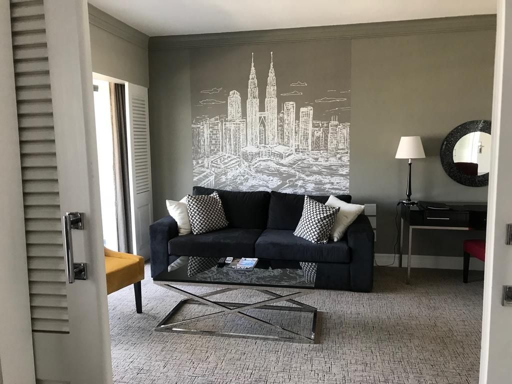 Ein Wohnzimmer mit Stadtansicht-Wandgemälde, einem schwarzen Sofa mit gemusterten Kissen, einem Couchtisch, einer Lampe und einem Spiegel an der Wand.