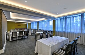 A hotel dining area with multiple tables and chairs, large windows with curtains, and a carpeted floor. - Capetonian Hotel (Cape Town)