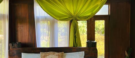 Un dormitorio con cabecera de madera, cama con sábanas blancas, almohada decorativa y ventana con cortinas verdes.