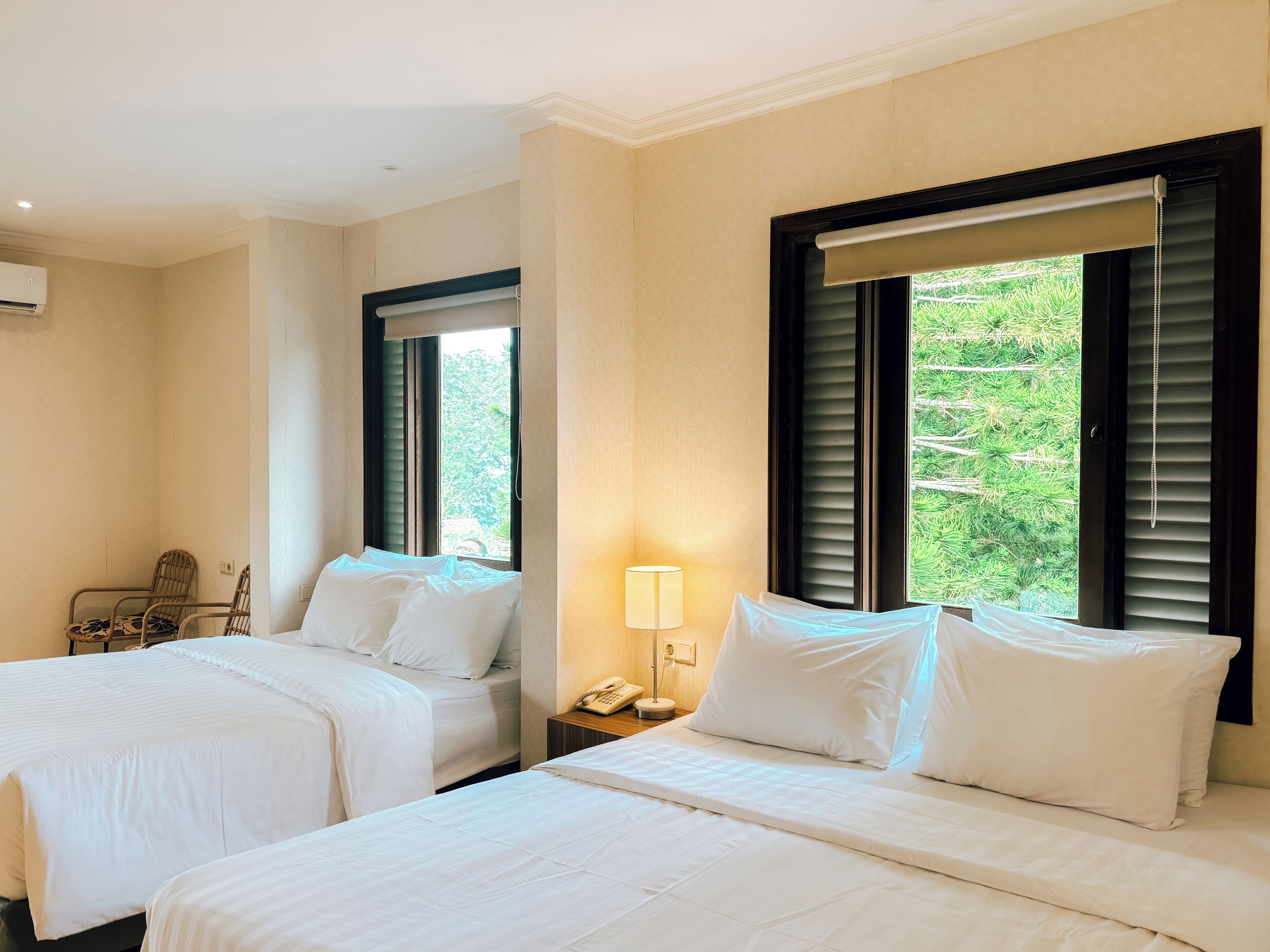 A hotel room with two beds, a chair, a bedside table with a lamp, and a window with blinds.