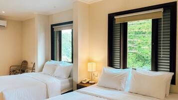 A hotel room with two beds, a chair, a bedside table with a lamp, and a window with blinds.