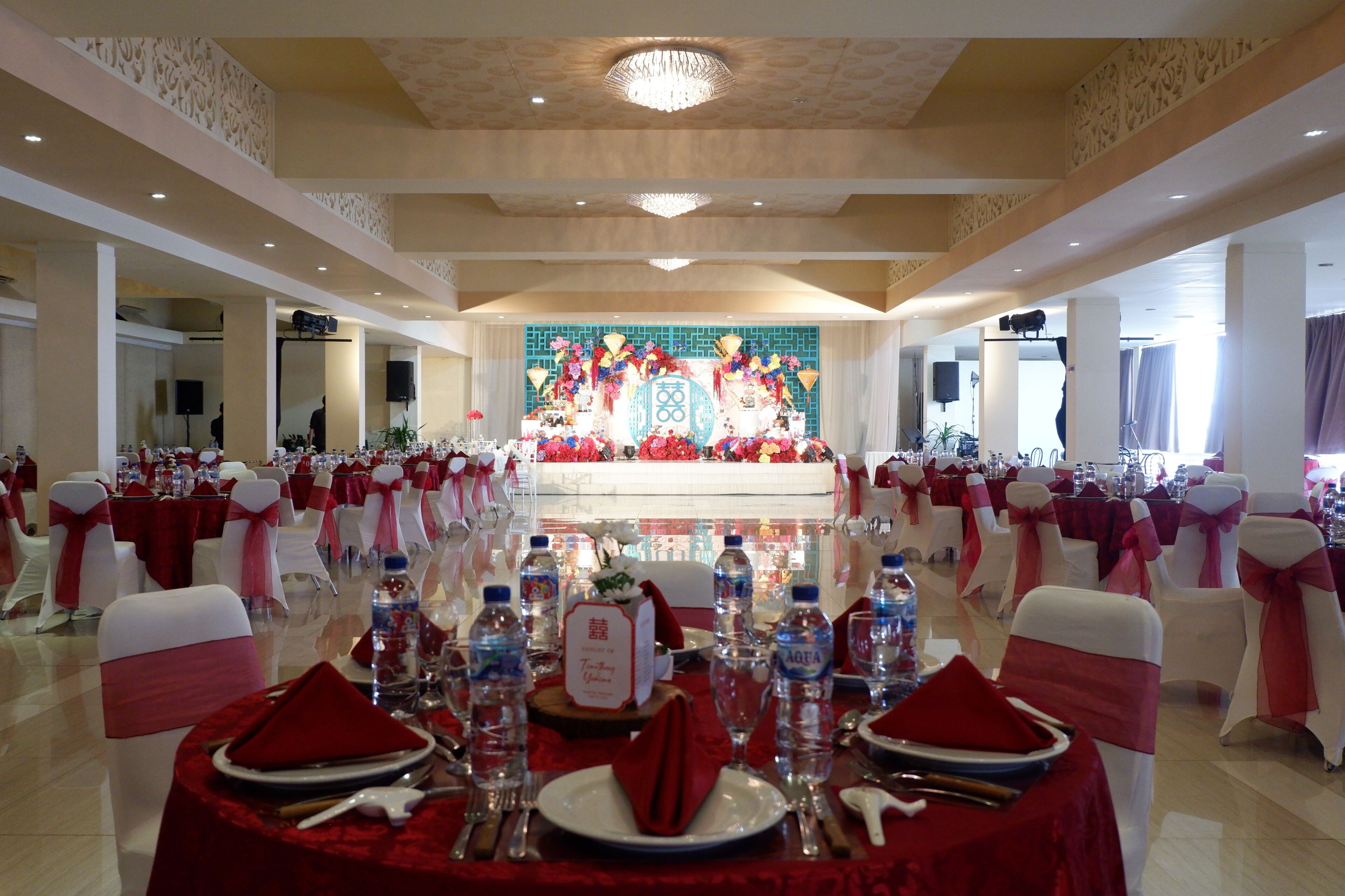 Un salón de banquetes con mesas redondas dispuestas para un evento formal, con un escenario central adornado con flores.